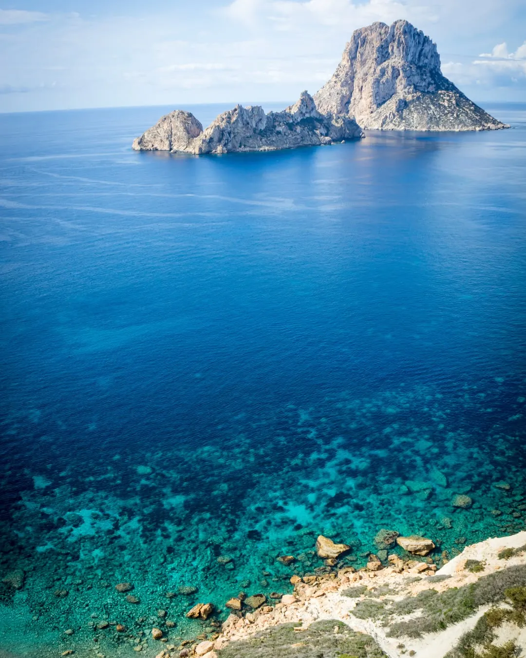 El islote de Es Vedrà visto desde la costa de Ibiza, con el mar azul profundo en primer plano y una franja de agua turquesa junto a la orilla.